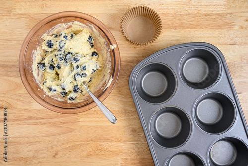 Muffin batter with fresh blueberries in a glass mixing bowl on wood counter, jumbo muffin tin with paper liners, baking lemon blueberry muffins at home
