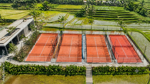 A captivating top-down aerial perspective highlights the vibrant contrast between four rust-orange padel courts, a modern concrete service pavilion, and the surrounding lush rice terraces.