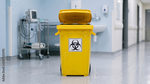 Yellow biohazard waste bin standing in a sterile hospital corridor with medical equipment background