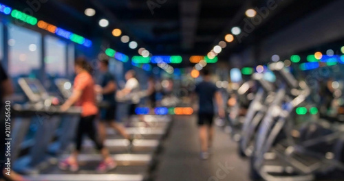A blurred view of a modern gym with people using treadmills and elliptical machines. Vibrant lights and a city view are visible through large windows.