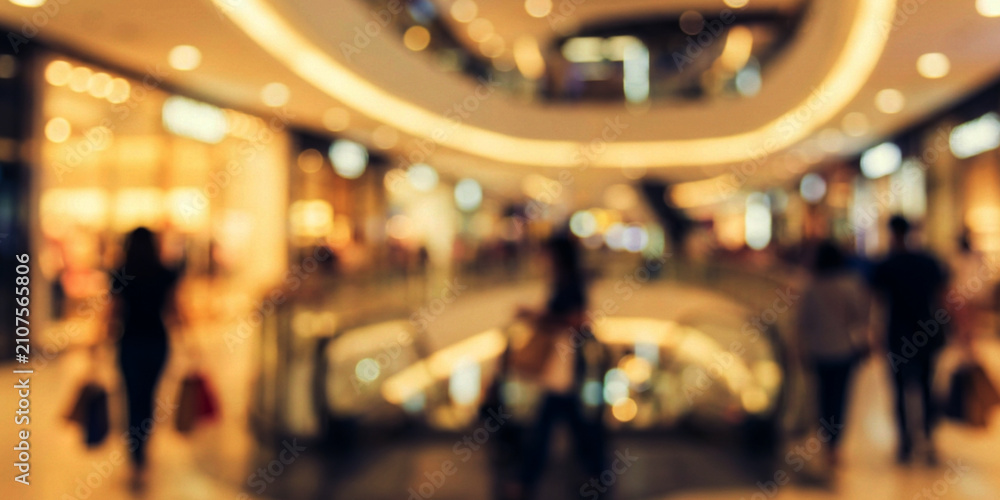 Fototapeta premium A blurred view of a shopping mall interior with escalators and shoppers, capturing a lively atmosphere in a retail environment.