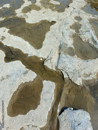Close-up of a natural mineral rock surface featuring white deposits, muddy water channels, and cracked textures. This abstract geological background showcases unique patterns formed by erosion