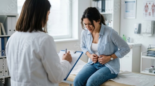 Young woman experiencing severe abdominal pain sits on examination table while professional female doctor takes notes during medical consultation in a bright clinic office to diagnose stomach issues