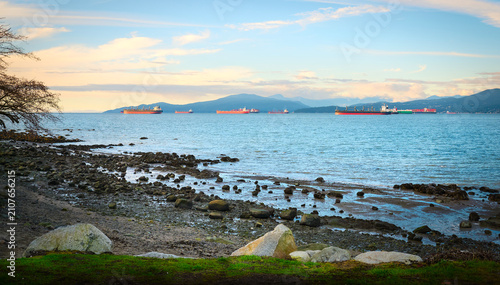 Cargo ships and bulk carriers anchored in English Bay viewed from a rocky shoreline with stones and sand under a cloudy blue sky at sunset, Vancouver, British Columbia, Canada.