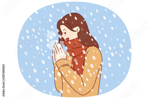 Woman enjoys winter weather, stands under falling snow flakes in sweater and scarf. Positive girl rejoices at onset of winter and approach of christmas holidays, blowing on hands to keep warm