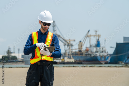 Photography Male marine engineer in hardhat and safety vest analyzing digital supply chain data on a tablet with cargo ship and seaport cranes in the background for maritime transport logistics