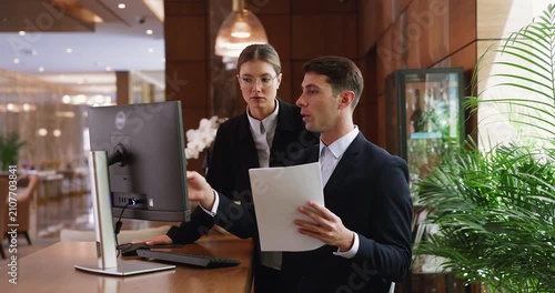 Two focused receptionists working together with hotel documentation behind registration desk, review papers, verifying booking details, check reservations, organize check-in forms for arriving guests