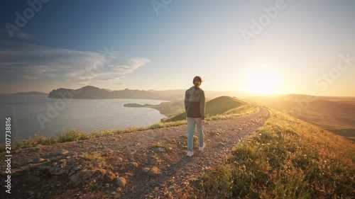 Young woman with a backpack hiking along a dirt path on a hill overlooking a beautiful sea bay at sunset. Cinematic steadicam follow shot footage for active lifestyle, summer travel and freedom video.