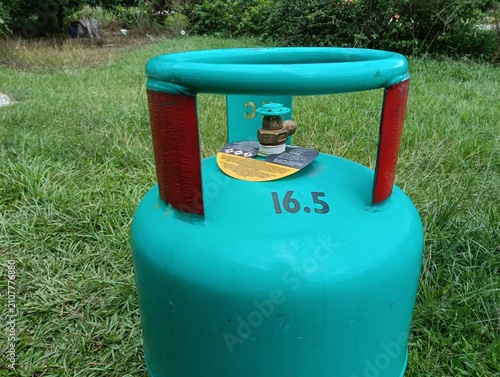 A green 16.5 kg LPG gas cylinder placed upright on a grassy field outdoors. The steel liquefied petroleum gas cylinder features a protective ring around the valve . in malaysia
