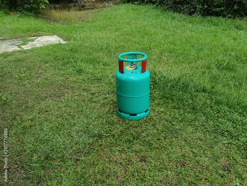A green 16.5 kg LPG gas cylinder placed upright on a grassy field outdoors. The steel liquefied petroleum gas cylinder features a protective ring around the valve . in malaysia