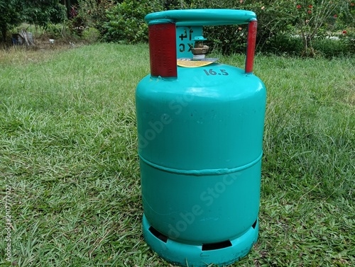 A green 16.5 kg LPG gas cylinder placed upright on a grassy field outdoors. The steel liquefied petroleum gas cylinder features a protective ring around the valve . in malaysia