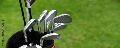 Golf clubs rest in a bag beside lush green grass. Metal heads gleam under bright sunlight. No people are visible in this shot. Ideal for sports, outdoor, or recreation themes