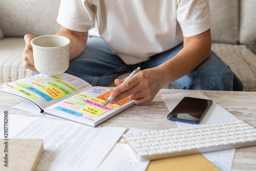 Person holding a pen and a mug, organizing appointments, expenses, and deadlines in a calendar planner book with colorful sticky notes, managing personal life and finances