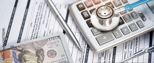 Medical expenses and budgeting. Stethoscope rests on calculator. Paperwork and cash show financial strain. Silver pen points to cost details. Image speaks of healthcare costs