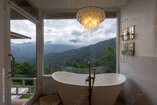 Freestanding bathtub with mountain view, large window framing misty forested hills, warm chandelier light, serene bathroom retreat with modern faucet and glass door