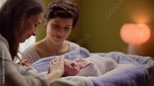 Two women interact lovingly with a swaddled newborn baby in a cozy indoor setting, soft lighting and warm colors creating a nurturing atmosphere