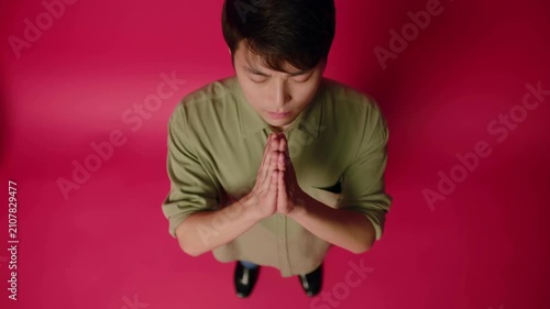 Asian male with black hair wearing green shirt stands with hands clasped in prayer position against a vibrant pink background, eyes closed in a moment of reflection