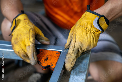 Worker wearing yellow leather glove holding metal frame and magnetic corner clamp, closeup of hands showing construction detail and focused craftsmanship