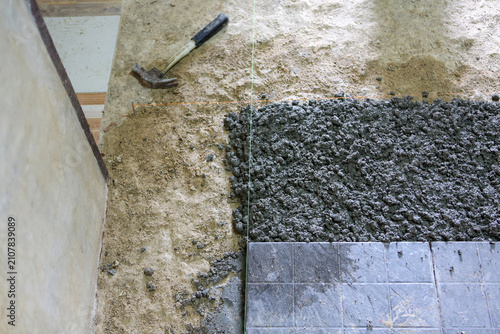 Wet cement floor being laid next to ceramic tile with hammer on rough subfloor, renovation scene showing texture and work in progress, gritty and practical atmosphere
