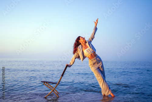 Photography Woman standing in shallow water holding a chair, reaching to the sky with a joyf