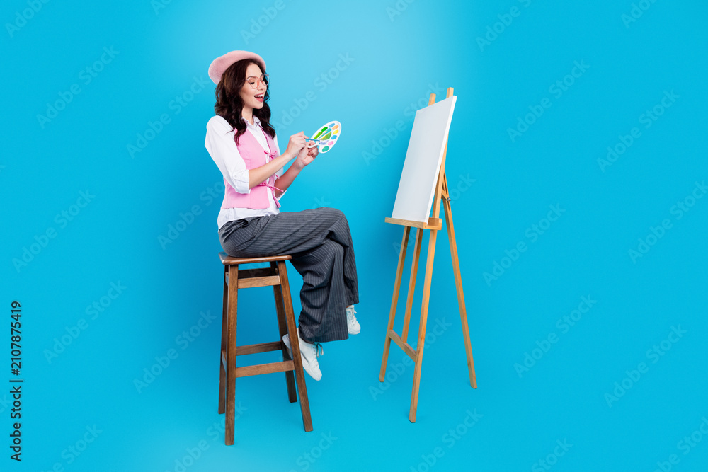 Fototapeta premium Young female artist painting on a blank canvas at an easel with palette and brush, wearing a pink beret and vest while sitting on a stool in a bright blue studio