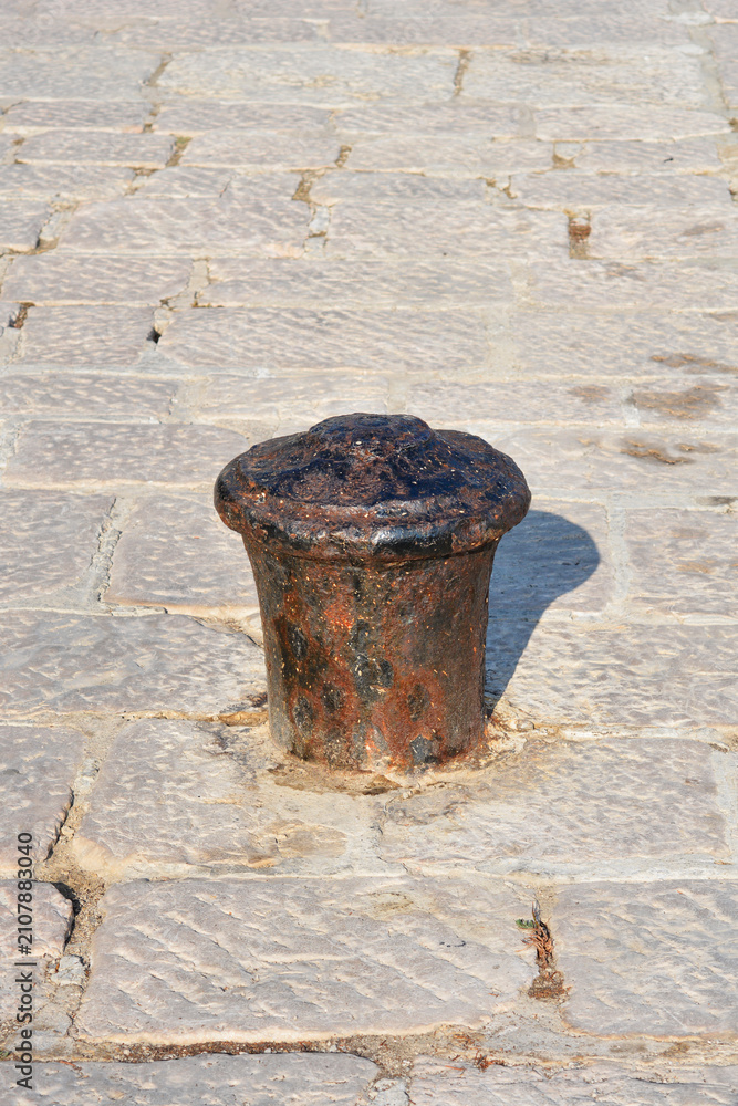 Fototapeta premium A vertical shot of an old rusty black iron mooring bollard mounted on a traditional stone paved street or dock surface under sunlight
