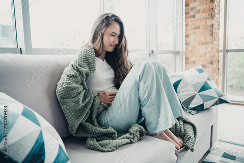 Obraz na płótnie Young woman sitting on couch with stomach pain wrapped in blanket holding abdome