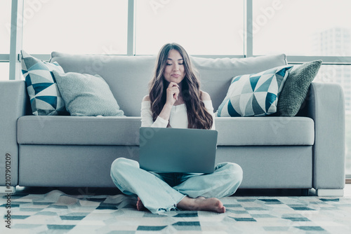 Young woman working on laptop at home sitting on floor in front of sofa, relaxed remote worker using computer in bright modern living room