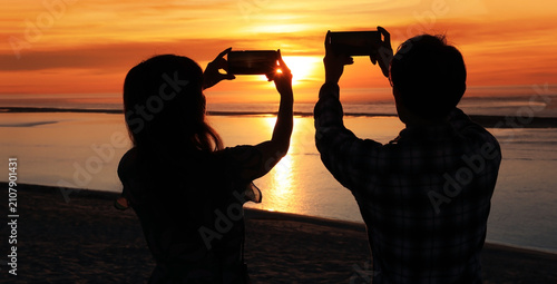 Silhouette of couple taking pictures of sunset  background on the sea beach with smartphones