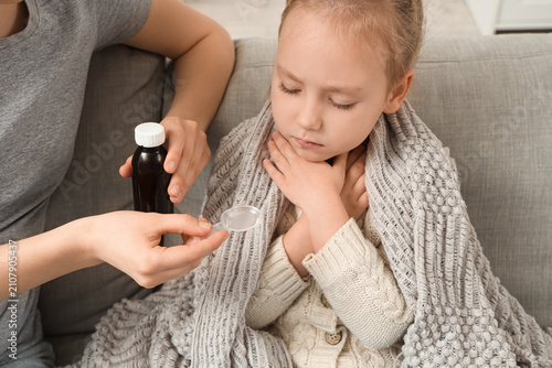Ill little girl taking cough syrup from her mother on sofa at home