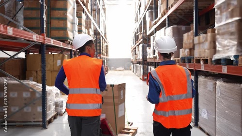 Two diverse female employees in hard hats and safety vests walking through a warehouse aisle, managing inventory and discussing logistics