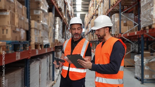 Two male logistics employees using a barcode scanner and a digital tablet for inventory management in a large distribution center aisle