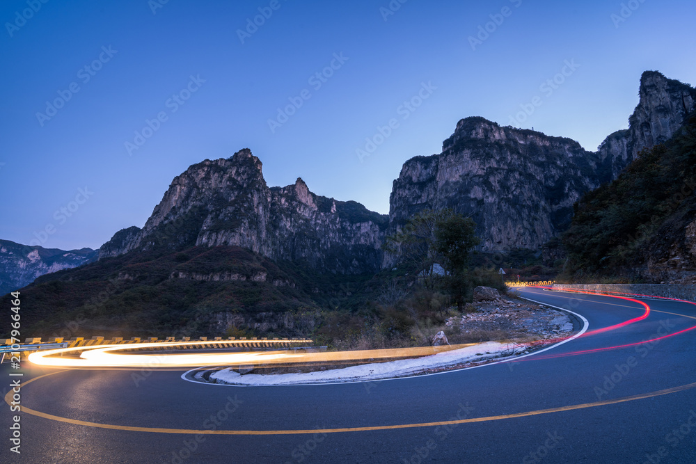 Obraz premium A long-exposure shot captures light trails from vehicles on a winding mountain road at twilight, surrounded by rocky peaks.