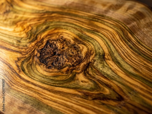 Close-up view of natural wood grain showing intricate swirling patterns, knots, and rich brown tones, ideal for organic textured backgrounds. Polished olive wood surface.