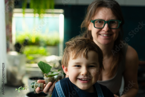 A happy mother and son explore a potted plant with leaves and flower in a sunny garden. The joyful gardener shares indoor greenery and seedling foliage with her playful boy in a warm moment.