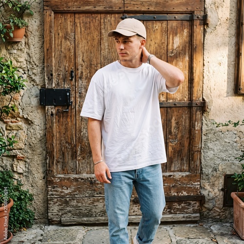 Young man wearing a white t-shirt and baseball cap stands in front of a rustic wooden door in an outdoor setting.
