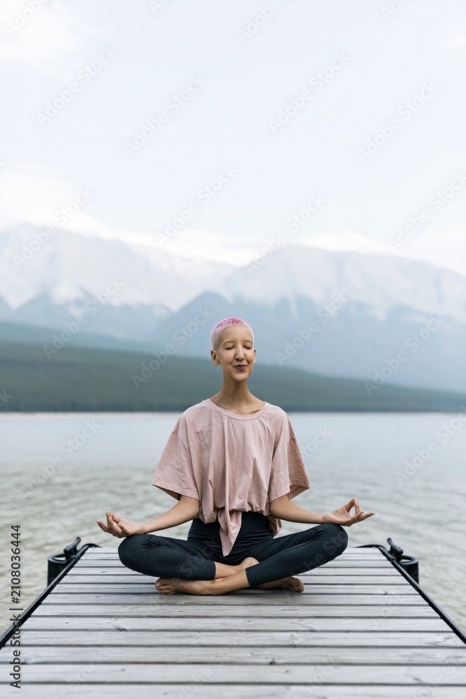 Fototapeta premium Smiling individual practicing seated meditation on a dock facing a mountain lake for mindfulness background and wellness banner with soft natural tones
