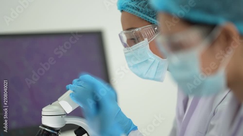 A masked scientist in cap and safety glasses handles a specimen slide at a microscope with purple cell tissue on the monitor.