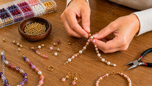 Close-up of hands making beaded jewelry on a wooden table. Artisan crafting handmade necklace with colorful beads and pliers. DIY hobby and small business concept