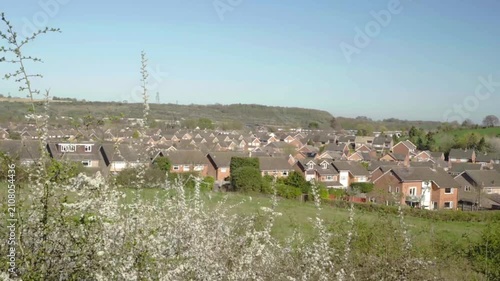 White-blossomed shrubs frame a grassy field leading to rows of suburban homes. Beyond them, open countryside and distant wind turbines complete the calm landscape.