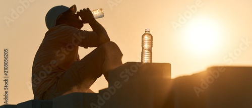 Construction worker drinking bottled water beside building materials at sunset, emphasizing hydration during extreme heat, suitable for occupational health, workplace safety and climate awareness proj
