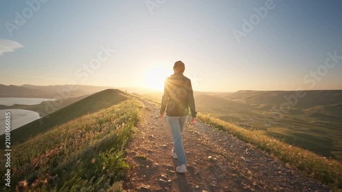 Young woman hiking along a dirt path on a hill overlooking a beautiful sea bay at sunset. Cinematic slow motion steadicam follow shot footage for active lifestyle, summer travel and freedom video.