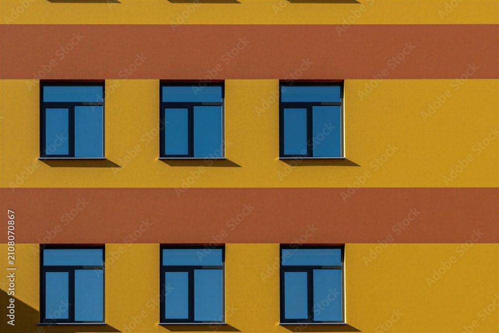 Obraz premium Geometric building facade with yellow and orange stripes. Six blue windows repeat in pattern. Modern architecture detail with clean lines and strong contrast. Minimalist exterior wall.
