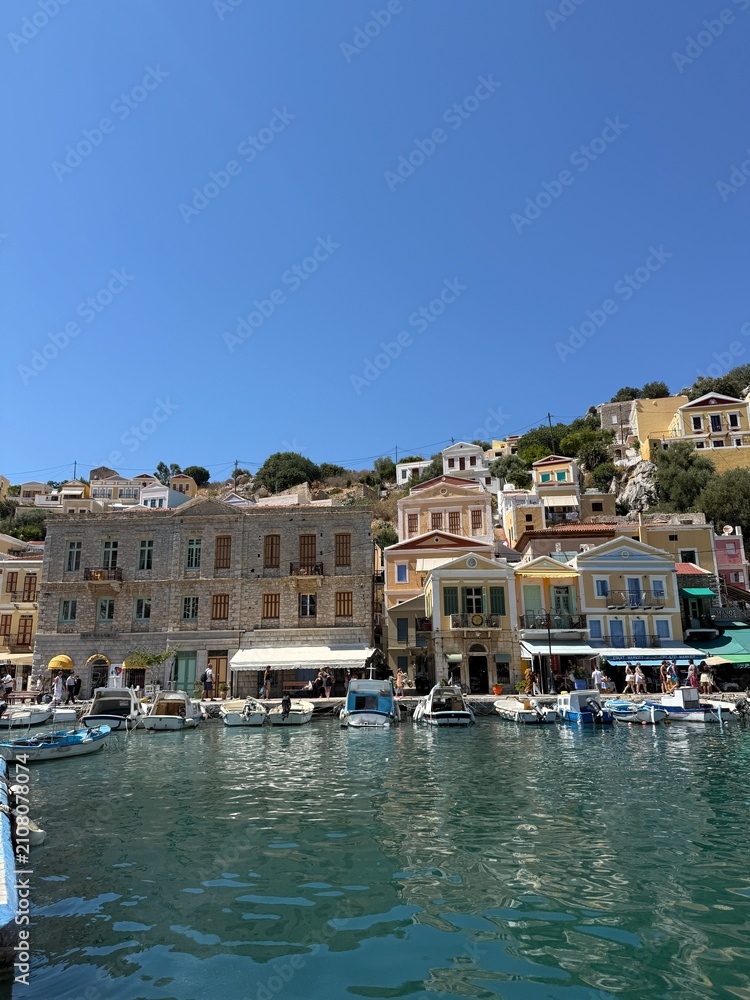 Obraz premium Colorful Symi harbor houses by the sea, turquoise Aegean water, traditional Greek island architecture, bright summer sunlight, peaceful coastal view