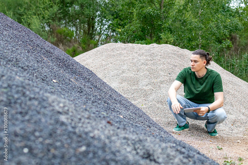Obraz na płótnie Middle aged man examines a pile of dark crushed stone while holding a tablet during an outdoor site visit