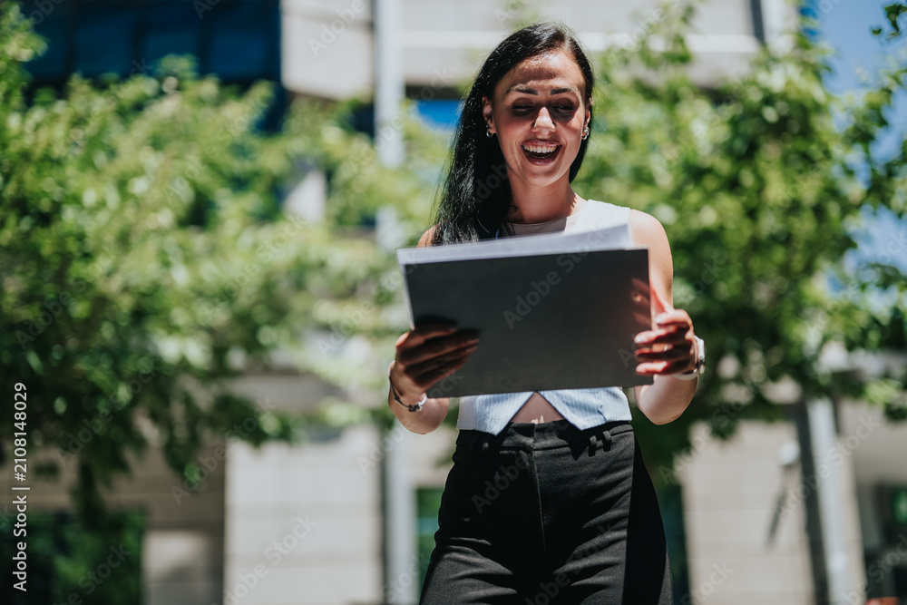 Fototapeta premium Young professional woman holds a laptop and smiles during an outdoor work meeting. The same woman appears engaged and happy while using a device in a sunny urban setting.