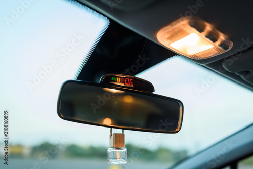 Rearview mirror with parking sensor display and air freshener inside car. Closeup view of vehicle's interior ceiling area with rear view mirror, digital parking indicator, and hanging perfume bottle
