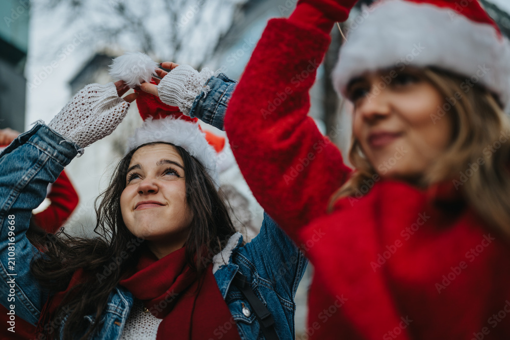 Fototapeta premium Two young women wear Santa hats and smile while adjusting their festive headwear. They stand outdoors in winter clothing enjoying a cheerful holiday moment.