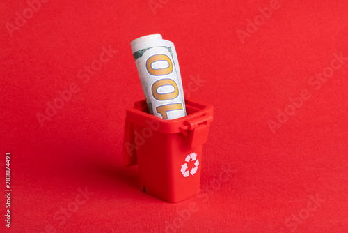 Rolled hundred dollar bill placed in a small red recycling bin on a solid red background, symbolizing financial waste or sustainability concepts.