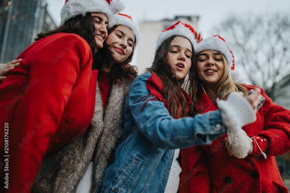 Fototapeta premium Four girls wearing Santa hats and winter coats pose closely while taking a selfie. They smile and celebrate outdoors in a festive, casual holiday moment.